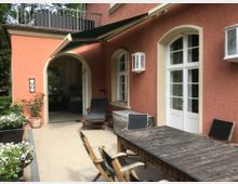 Terrasse eines Hauses mit roten Wänden und weißen Fenstern sowie Türen mit Rundbögen. Im Vordergrund stehen ein Holztisch und Stühle, daneben Blumentöpfe mit weißen Blüten; eine Markise spendet Schatten.