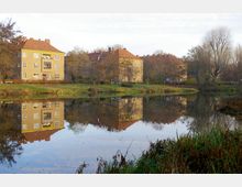 Mehrfamilienhäuser mit gelber Fassade und roten Ziegeldächern am Ufer eines ruhigen Teichs, in dem sich die Gebäude und umliegenden kahlen Bäume spiegeln. Der Vordergrund zeigt Gräser und Büsche entlang des Wassers.