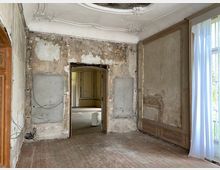 Ein leerer, renovierungsbedürftiger Raum in einem historischen Gebäude mit abblätterndem Putz an den Wänden, einem Holzboden und Stuckverzierungen an der Decke. Große Fenster mit teilweise durchsichtigen Vorhängen lassen Tageslicht herein, während eine offene Tür in einen angrenzenden Raum führt.