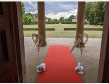 Blick von einem Eingang mit zwei Säulen auf eine Rasenfläche mit Kiesweg und Büschen, geschmückt mit einem roten Teppich und dekorativen, herzförmigen Metallstelen. Im Hintergrund stehen Gebäude und Bäume unter einem wolkigen Himmel.