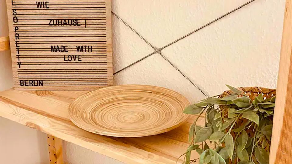 Holzregal an einer hellen Wand mit Querdrähten, darauf ein rundes Holztablett, Prospekte und eine hängende Grünpflanze. Links lehnt ein gerilltes Schild mit aufgedrucktem Text am Regal.