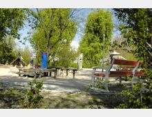 Ein Spielplatz in einer parkähnlichen Umgebung mit Bäumen im Hintergrund, einem kletterbaren Holzhaus, einem blauen Wasserspender und mehreren hölzernen Sitzbänken. Im Vordergrund steht eine rot-weiße Sitzbank, während Kinder im Bereich des Holzhauses spielen.