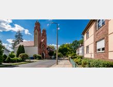 Straßenszene mit einer Kirche aus rotem Backstein, die durch zwei Türme mit spitzen grünen Dächern hervorsticht, umgeben von Bäumen und Grünflächen. Rechts daneben befinden sich helle Wohnhäuser mit Klinkerfassaden, und im Vordergrund verläuft ein Gehweg entlang eines begrünten Vorgartens.