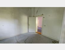 Leerer Raum mit weißen Wänden und abgenutztem grauem Bodenbelag. Eine leicht geöffnete Tür führt in einen angrenzenden Raum mit blassem rosa Anstrich und sichtbarem blauen Teppichboden.