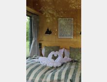 Innenansicht eines gemütlichen Schlafbereichs mit Doppelbett, gestreifter Decke und dekorativen Handtüchern in Herzform, flankiert von Kissen. An der Holzpaneelwand hängen zwei schwarze Wandlampen und ein typografisches Poster; links befindet sich ein großes Fenster mit grauen Vorhängen und Blick ins Grüne.