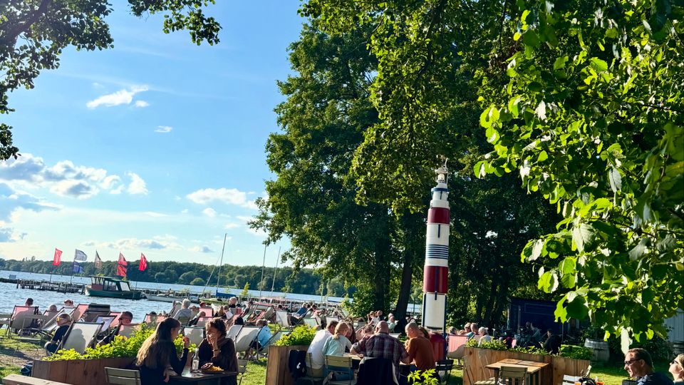 Ein Biergarten am Ufer eines Sees mit Blick auf Segelboote und rote Flaggen; im Vordergrund stehen Holztische und Stühle unter Bäumen, neben einem dekorativen rot-weißen Leuchtturm.