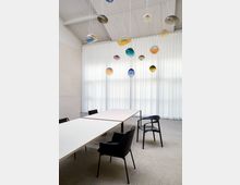 Moderner Besprechungsraum mit zwei rechteckigen Tischen, umgeben von schwarzen Stühlen. An der Decke hängen bunte, gläserne Pendelleuchten, im Hintergrund weiße, deckenhohe Vorhänge vor großen Fenstern.