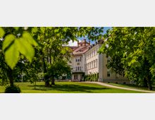Historisches Gebäude mit cremefarbener Fassade, großen Säulen und Terrakotta-Dach, umgeben von einer gepflegten Parklandschaft mit grünen Bäumen und Rasen. Ein geschwungener Weg führt durch die Grünfläche zum Eingang des Gebäudes.