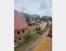 Ansicht eines verlassenen Gebäudekomplexes mit roten Ziegeldächern, teilweise eingestürztem Dach und überwucherten Teilen, umgeben von Bäumen. Im Vordergrund verläuft ein gepflasterter Weg entlang der Gebäude.