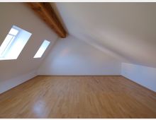 Dachgeschossraum mit weißen Wänden, einem schrägen Dach, sichtbaren Holzdeckenbalken und drei rechteckigen Dachfenstern auf einer Seite. Der Boden besteht aus hellem Holzparkett.