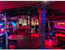 Innenraum einer Bar oder eines Clubs mit bunten LED- und Spotlichtern, Bar-Theke im Hintergrund und mehreren hohen Tischen mit Barhockern. Sichtbar sind eine Diskokugel, Steinpfeiler, eine abgehängte Decke sowie Sitznischen entlang der Wand.