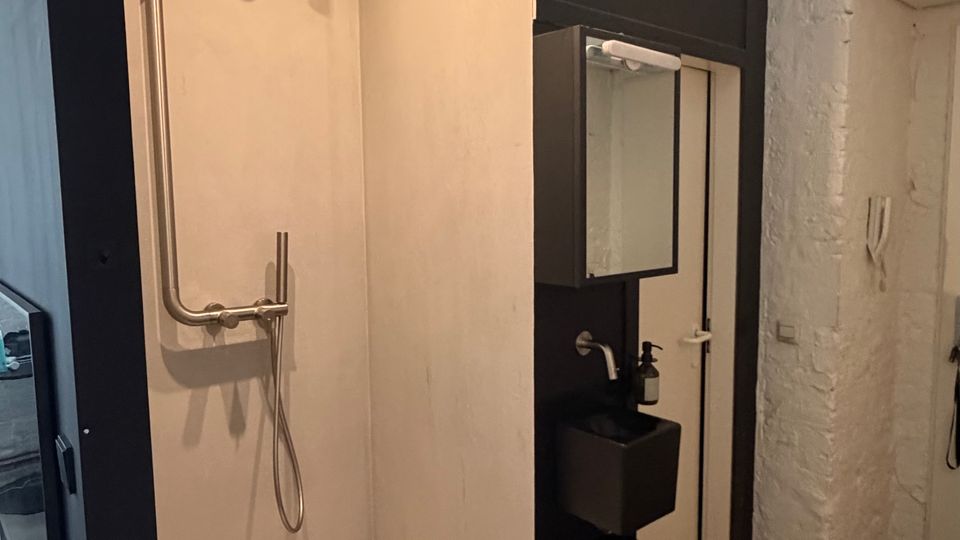 Modernes Badezimmer mit offener Duschkabine in schwarzer Rahmenkonstruktion, weißer Duschtasse und wandmontierter Armatur. Daneben ein kompakter Waschtischbereich mit Spiegel, schwarze Ablagen und eine weiße Ziegelwand unter einer gewölbten Decke.