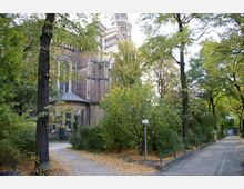 Backstein-Kirche mit neoromanischen Elementen, umgeben von dichtem Baumbestand und herbstlichem Laub auf Wegen und im Gebüsch. Im Vordergrund führt ein gepflasterter Weg entlang eines Zauns durch eine parkähnliche Umgebung.