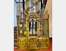 Historischer Aufzug mit reich verziertem, vergoldetem Metallgitter und kunstvollen Ornamenten in einem Gebäude. Im Hintergrund sind das Treppenhaus und hohe Fenster mit Rundbögen zu sehen.