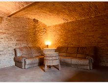 Gemütlicher Raum mit freiliegenden Ziegelwänden und einer gewölbten Decke. Zwei braune Ledersofas stehen um einen Tisch, der aus einem rustikalen Holzfass gefertigt ist, beleuchtet von einer warmen Lampe.