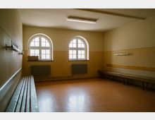 Ein einfacher Umkleideraum mit hellbraunen Wänden und einem beige-braunen Boden. Der Raum hat drei hohe Bogenfenster, zwei Holzbänke entlang der Wände und Hakenleisten zur Aufhängung, sowie Heizkörper unter den Fenstern.