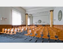 Innenraum eines Konferenz- oder Versammlungsraums mit mehreren Reihen aus Holzstühlen auf blauem Teppichboden. Der Raum ist geprägt von hohen Wänden mit holzvertäfeltem unteren Bereich, verzierten Decken, großen Fenstern mit vertikalen Jalousien und dekorativen Säulen aus gelbem Marmor.