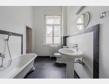 Helles Badezimmer mit freistehender Badewanne, Waschbecken auf Säule und WC, weiß gefliest mit dunkler Fliesenbordüre und dunklem Boden. Ein großes Fenster mit Heizkörper darunter sowie ein runder Wandspiegel und eine Leuchte ergänzen den Raum.