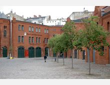 Ziegelstein-Gebäudekomplex mit Rundbogenfenstern und Toren, beschriftet mit "Wasch- und Umkleideräume" und "Lastauto-Garage", umgeben von einem gepflasterten Platz mit Bäumen und einigen Passanten. Im Hintergrund sind moderne Gebäude sichtbar.