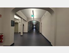 Langer, weiß gestrichener Innenflur in einem öffentlichen Gebäude mit Rundbögen, dunkelgrauem Boden und Deckenleuchten. In der Mitte führen gläserne Doppeltüren weiter in den Korridor, darüber sind grüne Notausgangsschilder angebracht.