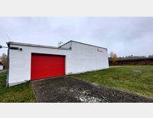 Ein einstöckiges weißes Gebäude mit flachem Dach und einem großen roten Rolltor an der linken Seite. Vor dem Gebäude befinden sich eine gepflasterte Fläche und eine Rasenfläche, im Hintergrund sind Bäume und ein bewölkter Himmel zu sehen.