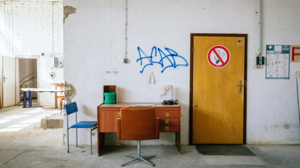 Ein leerer Raum in einem heruntergekommenen Gebäude mit einem weißen, teilweise beschädigten Putz an den Wänden. Im Vordergrund steht ein Holztisch mit einem braunen Stuhl und daneben ein blauer Stuhl; an der Wand befindet sich eine graue Tür mit einem "Rauchen verboten"-Schild darüber.