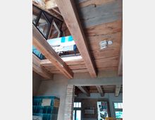 Innenraum mit offener Holzbalkendecke und sichtbaren Ziegelwänden. Im Vordergrund steht ein grün glasierter Kachelofen, im Hintergrund sind Fenster, eine Pinnwand und ein Kühlschrank sichtbar.