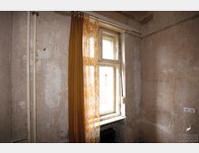 Ein kleiner, schlichter Raum mit unverputzten, verwitterten Wänden, einem alten Fenster mit gelbem Vorhang und einem Heizkörper darunter. Links sind zwei freiliegende Rohre sichtbar, und an der Wand befindet sich eine Steckdose.