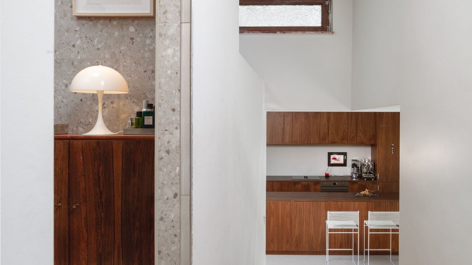 Minimalist interior hallway leading to a compact kitchen with dark wood cabinets, a narrow frosted window, and an exposed concrete ceiling. At left, an alcove with terrazzo walls contains a wooden cabinet topped with a white dome lamp and a framed picture.