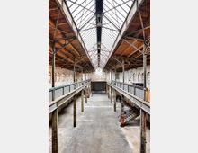 Alte Industriehalle mit hohen Decken, einem Dach aus Glas und Metallträgern, sowie sichtbaren Stahlstützen und Holzelementen. Der Raum verfügt über zwei offene Galerien an den Seiten und eine große, zentrale Freifläche aus Beton.