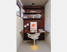Small built-in home office nook with a dark wood desk and wall-mounted shelving, set between white walls under an exposed concrete ceiling. A window on the left brings in daylight, and the desk holds a globe lamp, stacked books, and framed artwork, with a white pedestal chair in front.