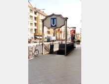 U-Bahn-Eingang mit einem historischen Schild mit großem blauen "U" über der Treppe, die zum unterirdischen Bahnhof führt. Im Hintergrund sind eine belebte Straße mit Autos, Geschäftsgebäuden und Werbeplakaten zu sehen.