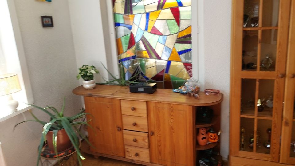 Wohnlicher Innenraum mit einem großen, bunten Buntglasfenster an der Wand und einem niedrigen Holzsideboard davor. Neben dem Sideboard stehen Zimmerpflanzen, rechts ist eine hohe Vitrine aus Holz mit Glastüren zu sehen.