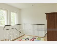 Innenansicht eines hellen Treppenhausabsatzes mit weiß gestrichenem Geländer und modernen runden Verzierungen. Links befinden sich zwei große Fenster, die Tageslicht hereinlassen, und der Boden ist mit einem Teppich mit buntem, floralem Muster ausgelegt.