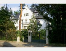 Großes weißes Herrenhaus im klassischen Stil mit verzierten Giebeldächern, mehreren Fensterreihen und einem kleinen Balkon; umgeben von Bäumen und dichtem Grün. Im Vordergrund steht eine schmiedeeiserne, von zwei Säulen gesäumte Eingangstor-Anlage, die von einer Hecke und einem Zaun flankiert wird.