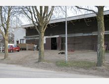 Ein einstöckiges Gebäude mit dunkler Holzfassade, großen doppelflügeligen Türen und einem flachen Dach, umgeben von kahlen Bäumen. Im Vordergrund sind ein geparkter roter Kleinwagen, eine sandige Fläche und ein Hund zu sehen.