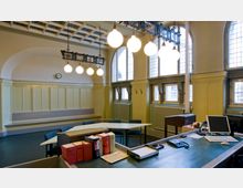 Innenraum eines historischen Büros oder Verwaltungsgebäudes mit hohen Decken, großen Rundbogenfenstern mit Sprossen, einer gelblichen Wandverkleidung und einem dekorativen Kronleuchter mit kugelförmigen Lampen. Im Vordergrund stehen ein Schreibtisch mit Computer und Büchern sowie ein Konferenztisch mit Stühlen.