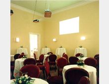 Ein Veranstaltungsraum mit hellgelben Wänden, roten Polsterstühlen und runden, weiß gedeckten Tischen, die mit Blumenarrangements dekoriert sind. An den Wänden befinden sich wandmontierte Lampen, und im Raum hängen Projektoren von der Decke.