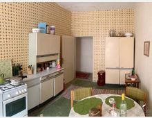 Küche im Retro-Stil, mit gelb-gemusterter Tapete, einer weißen Küchenzeile und einem älteren Gasherd. Im Raum befinden sich ein runder Esstisch mit grünen Platzdeckchen, Retro-Möbel und verschiedene Haushaltsgegenstände.