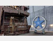 Industriehalle mit einer massiven, rostigen Maschine auf der linken Seite und einem großen, blauen Stern mit einem „B“ in der Mitte auf der rechten Seite. Dahinter befinden sich graue Betonwände und helle Beleuchtungselemente.