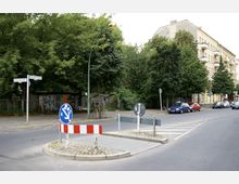Straßenkreuzung mit Verkehrsinseln und Schildern, umgeben von Bäumen und Mehrfamilienhäusern. Im Hintergrund sind geparkte Autos, eine Hausfassade und eine eingezäunte Grünfläche zu sehen.