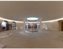 Runder Innenraum mit glattem, hellbeigem Steinboden und zentraler Deckenöffnung mit diffusem Licht. Um die Mitte sind Säulen angeordnet, und an den Wänden stehen weiße Silhouetten von Tieren wie Pferden, einer Kuh und einem Schaf; darüber hinaus befinden sich einige bogenförmige Eingänge mit Glas- und Steinelementen.