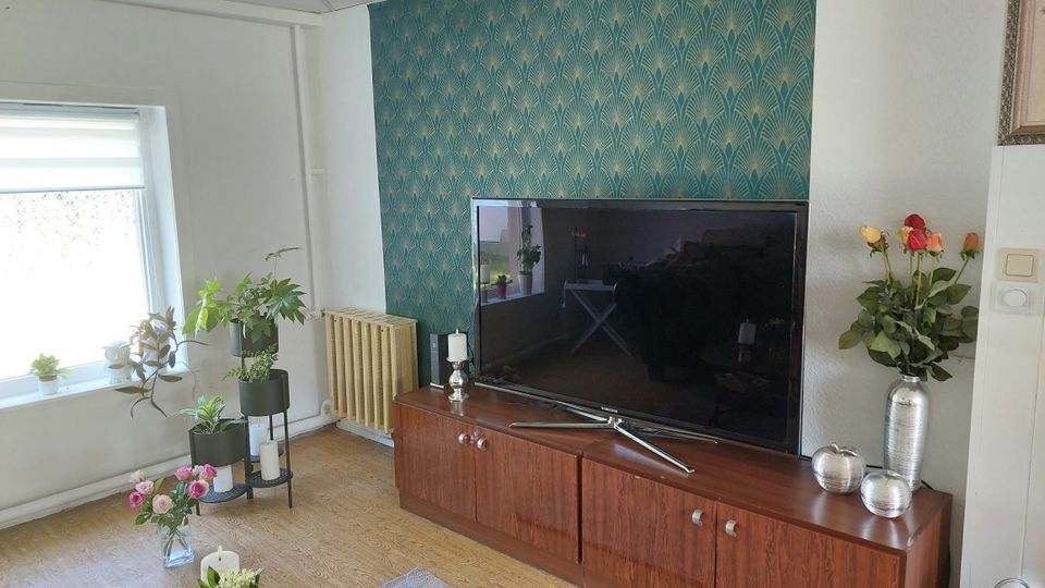 Wohnzimmer-Ecke mit großem Flachbildfernseher auf einem niedrigen Holz-Sideboard vor einer türkis gemusterten Akzentwand. Links ein Fenster mit Rollo, mehrere Zimmerpflanzen auf Ständern und ein Heizkörper; auf dem Sideboard stehen Kerzen und eine Vase mit Rosen.