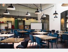 Ein modernes Café oder Veranstaltungsraum mit Holztischen und gepolsterten blauen Stühlen, darüber hängen schwarze Hängeleuchten. Im Hintergrund stehen Regale mit Dekorobjekten sowie eine Leinwand vor einer grün gemusterten Wand.