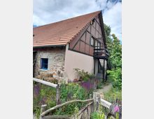 Fachwerkhaus mit Ziegeldach, seitlich mit einem kleinen Balkon und Außentreppe. Im Vordergrund ein Garten mit blühenden Pflanzen, darunter Lavendel, und ein rustikaler Holzzaun, umgeben von Bäumen und Natur.