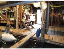 Holzbauwerk mit Steg und Anlegestelle im Innenbereich, umgeben von rustikalen Holzbalken und Regalen mit Alltagsgegenständen. Im Vordergrund schwimmt ein kleines Schlauchboot im Wasser; Laternen und Ketten sind an den Pfosten befestigt.