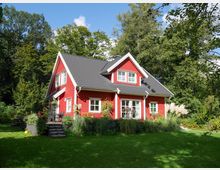 Ein rotes Holzhaus mit schwarzem Satteldach und weißen Fenstereinfassungen, umgeben von einem üppigen Garten mit Pflanzen, Bäumen und Rasen. Das Haus hat eine Terrasse mit Stufen und sitzt in einer ländlichen, grünen Umgebung.