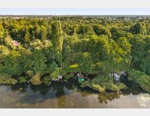 Luftaufnahme eines bewaldeten See- oder Flussufers mit dichtem Baumbestand, Schilf- und Seerosenflächen im flachen Wasser. Am Ufer liegen mehrere kleine Stege mit Booten, im Hintergrund erstreckt sich eine flache Landschaft mit vereinzelter Bebauung.