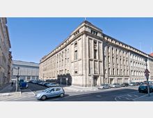 Eckgebäude mit massiver Steinfassade im Stil der klassischen Architektur, gekennzeichnet durch vertikale Fensterreihen und klare geometrische Linien. Im Vordergrund eine Straßenkreuzung mit parkenden Autos und Verkehrsschildern, bei klarem Himmel.