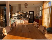 Esszimmer und offene Küche in einem sonnendurchfluteten Raum mit großen Sprossenfenstern und gelben Vorhängen. Der Raum enthält einen langen Holztisch mit gepolsterten Stühlen, eine Küchenzeile mit Edelstahl-Dunstabzugshaube sowie einen Kühlschrank und eine Spüle.